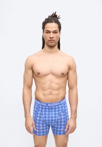 Homme en forme avec des dreadlocks portant des boxers bleus à motifs avec une ceinture à logo Björn Borg, debout devant un fond blanc uni.