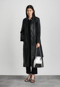 Black leather trench coat with button closure, worn over a black outfit. Paired with a silver fringe-accented bag and black strappy sandals.