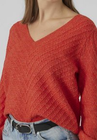 Teksturowany czerwony sweter z dekoltem w kształcie litery V, z wzorem w zygzaki. Noszony na jasnoniebieskich dżinsach i czarnym pasku. Swobodny krój, szczegóły w prążki.
