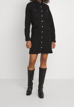 Personne portant une robe noire à manches longues boutonnée avec poches poitrine et bottes en cuir noires montant jusqu'aux genoux, sur un fond uni.