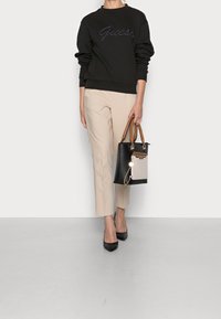 Sort sweatshirt med broderet "Guess"-logo, beige tætsiddende bukser, sorte stiletter og en sort håndtaske med brune håndtag og cremefarvet detalje.