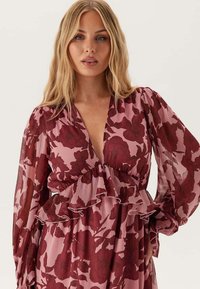 Blond kvinna klädd i en rosa klänning med stora mörkröda blommönster och volanger, stående mot en enfärgad bakgrund.