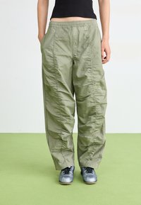 Licht olijfgroene, loszittende broek van lichtgewicht materiaal, met verticale naden en een elastische tailleband. Geschreven met grijze schoenen.