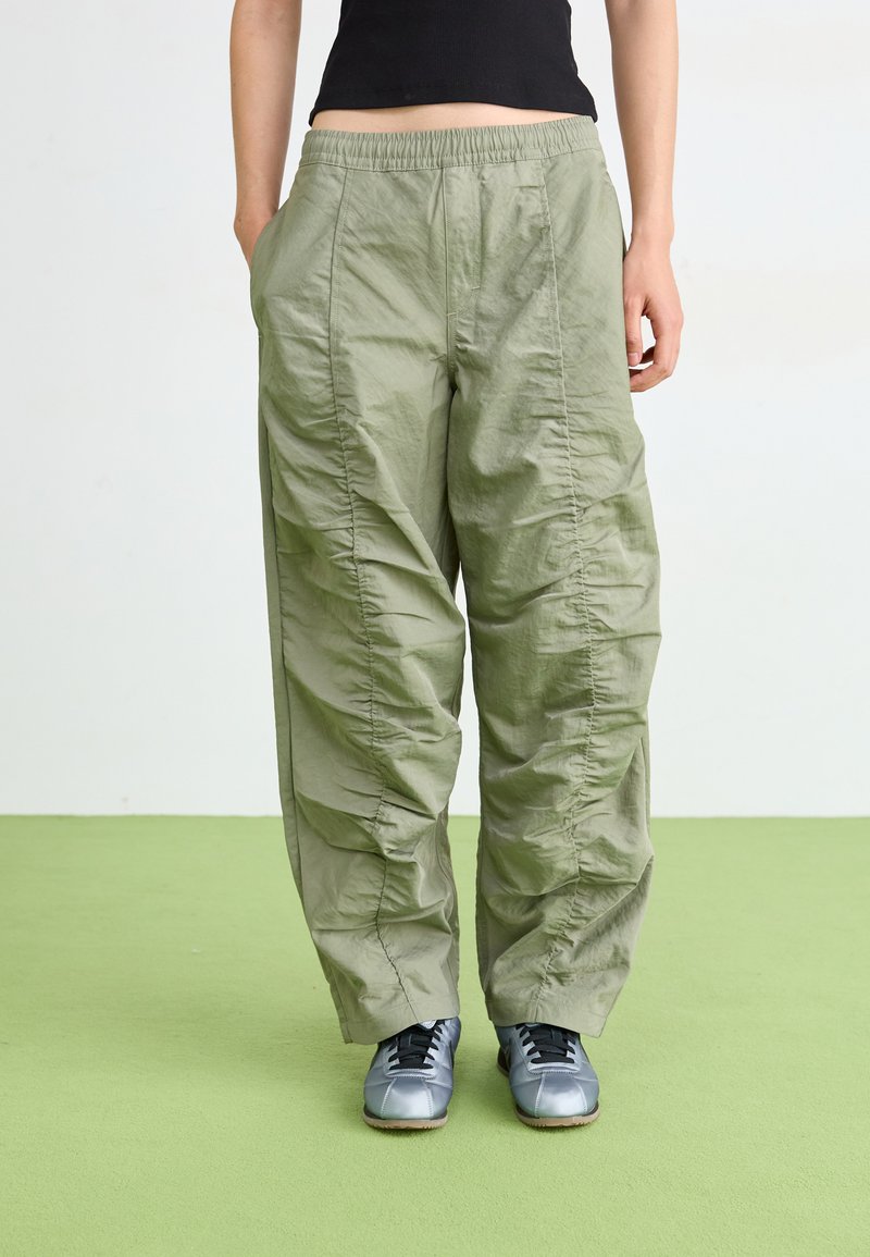Licht olijfgroene, loszittende broek van lichtgewicht materiaal, met verticale naden en een elastische tailleband. Geschreven met grijze schoenen.