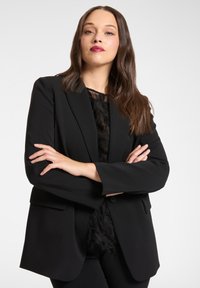 Blazer nero con vestibilità sartoriale, caratterizzato da rever a lancia e chiusura con un solo bottone. Indossato sopra una maglia nera testurizzata, con maniche lunghe.