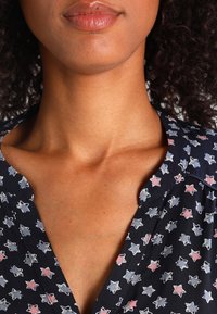 Camicia di tessuto scuro con un motivo a stelle in rosa e blu chiaro. Il colletto è aperto, mettendo in risalto il décolleté e l'area delle spalle.