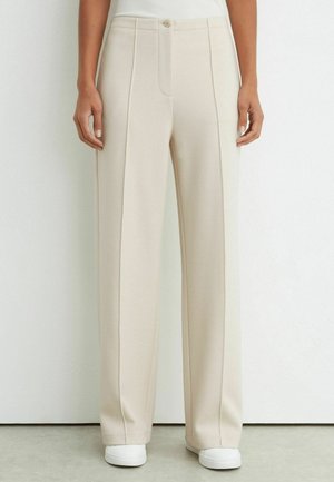 Woman wearing high-waist, cream-colored wide-leg trousers with front seams and white sneakers against a plain white wall.