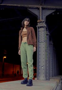 Brun zip-up jacka, brun randig crop top, gröna avslappnade byxor och svarta chunky boots. Modellen står under en stålkonstruktion, urban miljö.