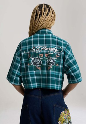 Osoba z długimi blond warkoczami nosi zieloną kraciastą krótka koszulę z napisami "Ed Hardy" i wzorami róż na plecach, w połączeniu z ciemnoniebieskimi spodniami.