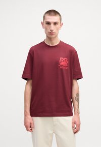 Burgundy cotton t-shirt featuring a small red logo on the chest, crew neckline, and short sleeves, paired with light-colored pants.