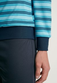 Blauwe en witte gestreepte lange mouwen shirt met marineblauwe manchetten, gecombineerd met donkergrijze broek. De stof is zacht en glad. Detailtag zichtbaar.