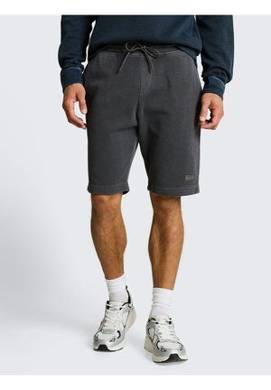 Man draagt donkergrijze shorts met trekkoord, witte sportsokken en witte en grijze hardloopschoenen, staand tegen een effen achtergrond.