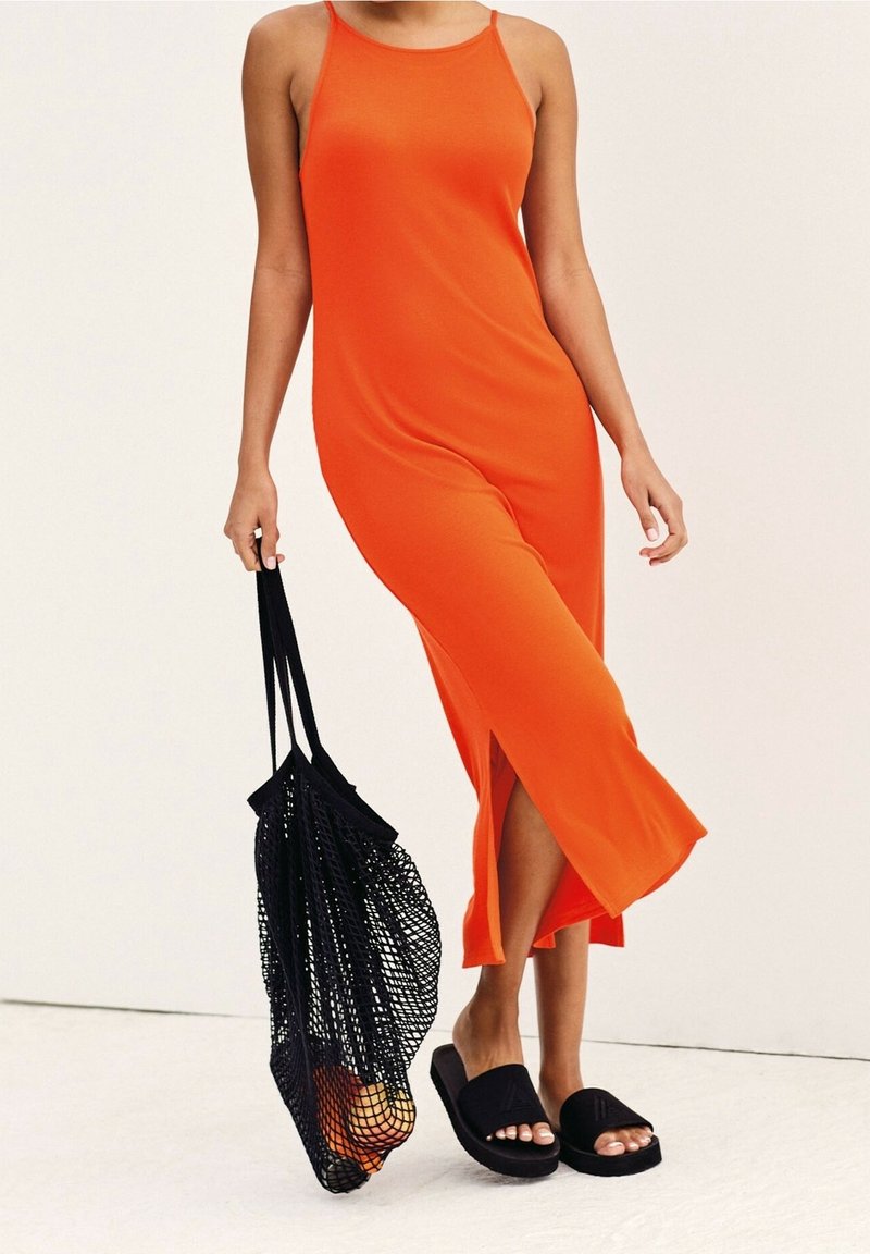 Robe orange sans manches avec fentes latérales, réalisée en un tissu lisse. Associée à des sandales slides noires et à un sac fourre-tout en filet noir.