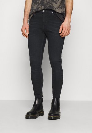 Mann trägt schlanke schwarze Jeans und schwarze Leder-Stiefeletten, steht vor weißem Hintergrund.