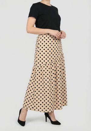 Femme portant une jupe midi beige à pois noirs, un haut noir à manches courtes et des talons hauts noirs, debout devant un fond uni.