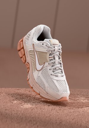 Hvid og beige Nike Vomero 5 sneaker med mesh og gummisål, placeret på en struktureret brun overflade mod en neutral baggrund.