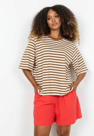 Kvinde med krøllet hår iført en brun- og hvidstribet t-shirt og lyserøde shorts, stående med hænderne i lommerne mod en hvid baggrund.
