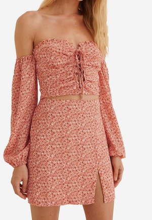 Frau trägt ein pinkes geblümtes Off-Shoulder-Crop-Top mit Schnürung vorne und passenden hoch taillierten Rock mit Seitenschlitz.