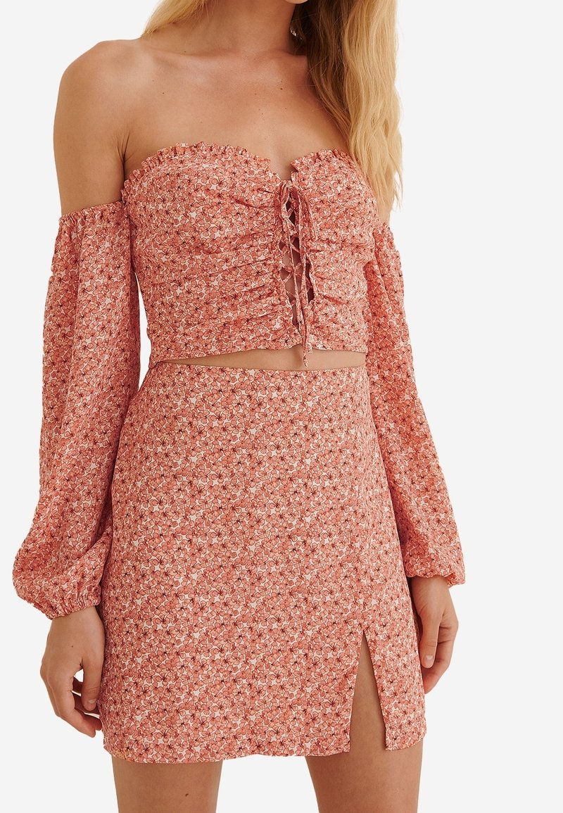 Femme portant un crop top fleuri rose à épaules dénudées avec un laçage devant et une jupe taille haute assortie avec une fente latérale.