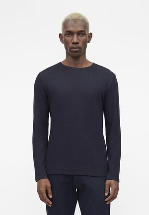 Uomo nero con capelli corti biondi che indossa una maglia aderente blu navy a coste e maniche lunghe, in piedi davanti a uno sfondo bianco semplice.