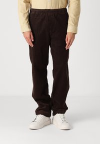 Bruna kordbyxor med elastisk midja, med rak passform och tillsammans med vita sneakers på en neutral bakgrund.
