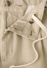 Pantalon beige avec fermeture par bouton et zip, montrant des boutonnières et un cordon blanc sur un tissu texturé.