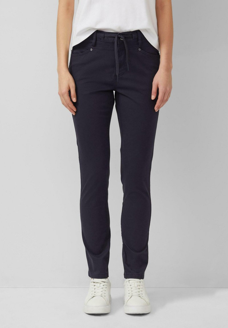 Vrouw draagt nauwsluitende donkerblauwe broek met trekkoord in de taille en witte sneakers, staand tegen een effen lichtgrijze achtergrond.