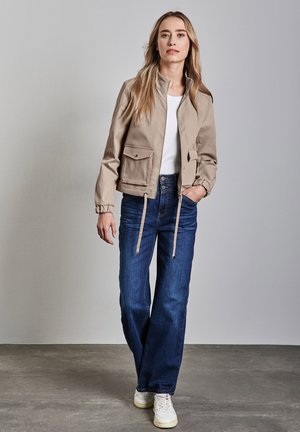 Veste cropped beige avec deux poches avant, poignets élastiques et détail à cordon, associée à un jean bleu taille haute et des baskets blanches.