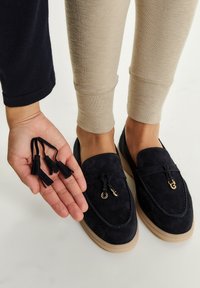 Loaferi din daim albastru marin cu o talpă deschisă la culoare, prezentând un accent decorativ din hardware auriu și ciucuri asortați ținuți într-o mână.