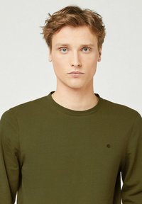 Giovane uomo con capelli castano chiaro che indossa una camicia a maniche lunghe verde oliva con un piccolo logo di un albero ricamato sul petto.
