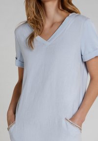 Lichtblauw V-hals top met opgerolde korte mouwen, voorzien van twee zakken versierd met een subtiele metalen rand. Glad, zacht textuur.