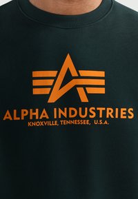 Sötétzöld pulóver narancssárga grafikával, amelyen az "A" betű és három csík látható, valamint a következő szöveg: "ALPHA INDUSTRIES, KNOXVILLE, TENNESSEE, U.S.A."