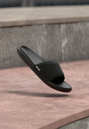 Schwarze Reef Slide-Sandale mit strukturiertem Fußbett, perforiertem oberen Riemen und "REEF"-Logo an der Seite, schwebend über einer Steinoberfläche gezeigt.