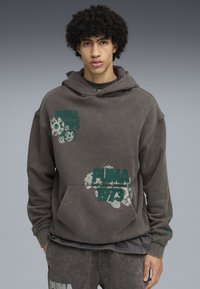 Felpa grigia oversize con accenti verdi del logo "PUMA", tasca anteriore e cappuccio con cordino. Realizzata in un morbido tessuto testurizzato con un look vissuto.
