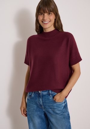 Femme aux cheveux bruns souriante, portant un pull bordeaux à manches courtes et un jean bleu, debout avec une main dans la poche devant un fond uni.