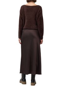 Personne portant un pull en peluche marron foncé, une jupe satinée longue assortie et des bottes noires épaisses, se tenant de dos.