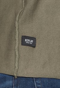 Tissu tricoté vert olive avec une étiquette noire visible portant l'inscription « REPLAY SINCE 1981 ». Les détails de couture et la texture sont bien visibles. Denim bleu foncé visible en dessous.