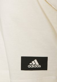 Krémové bavlněné tričko s černým obdélníkovým štítkem, na kterém je bílé logo Adidas. Výrazné švy podél lemu.