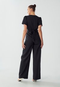Zwarte jumpsuit met korte mouwen, band rondom de taille, brede pijpen en een gladde textuur; heeft een sluiting aan de achterkant en een minimalistisch ontwerp.