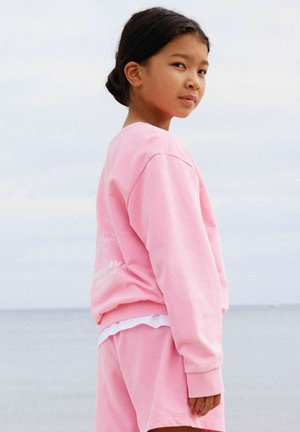 Meisje met donker haar in een paardenstaart, gekleed in een roze trui en korte broek, staand opzij bij rustig water onder een bewolkte hemel.