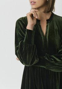 Grünes Samtkleid mit V-Ausschnitt, gerafften Puffärmeln und fließender Silhouette. Glatte Textur mit dezentem Glanz.