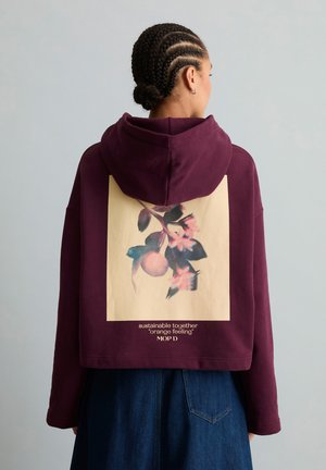 Person mit geflochtenem Haar, trägt einen bordeauxfarbenen kurzen Hoodie mit Blumenmuster und Text auf dem Rücken, kombiniert mit blauen Jeans.