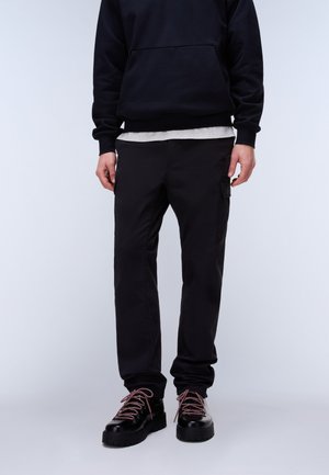 Bărbat purtând pantaloni cargo negri, bluză neagră peste o cămașă albă și pantofi negri cu șireturi roz, stând în picioare pe un fundal simplu.