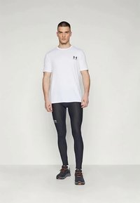 Vīrietis stāv, uzvilcis baltu īsām piedurknēm sporta kreklu, melnas kompresijas legins un melnus skriešanas apavus ar oranžiem akcentiem.