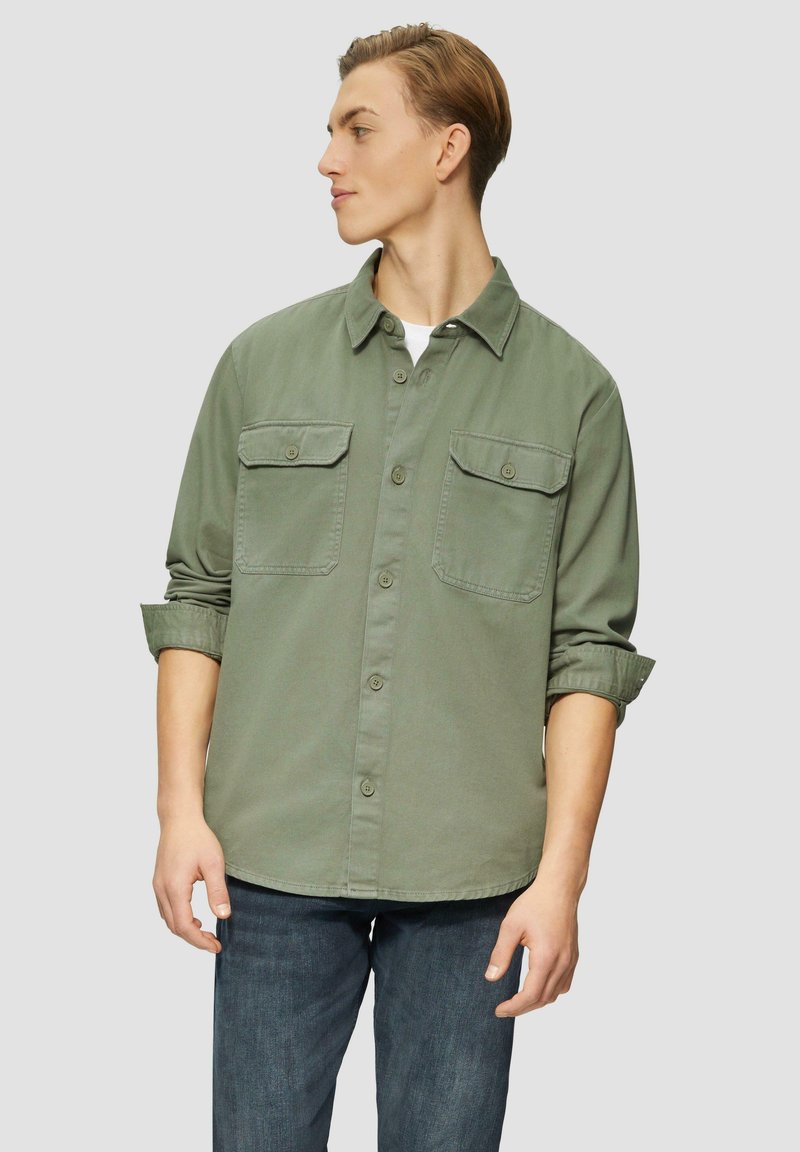 Button-up shirt in olijfgroen, met twee borstzakken, een neklap en opgerolde mouwen. De stof lijkt textuur te hebben en is zacht.