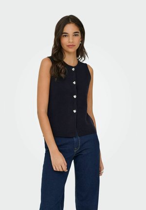 Mlada ženska z dolgimi temnimi lasmi, oblečena v črno brezrokavno bluzo s stiski in temno modre kavbojke, stoji ob enobarvni svetli ozadju.