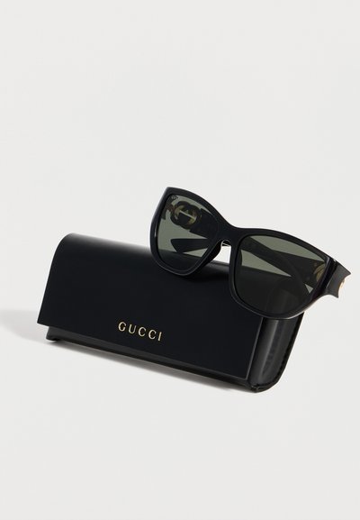 Μαύρα υπερμεγέθη γυαλιά ηλίου Gucci με σκούρους φακούς που ακουμπούν σε μαύρη δερμάτινη θήκη Gucci με χρυσό λογότυπο σε λευκό φόντο.