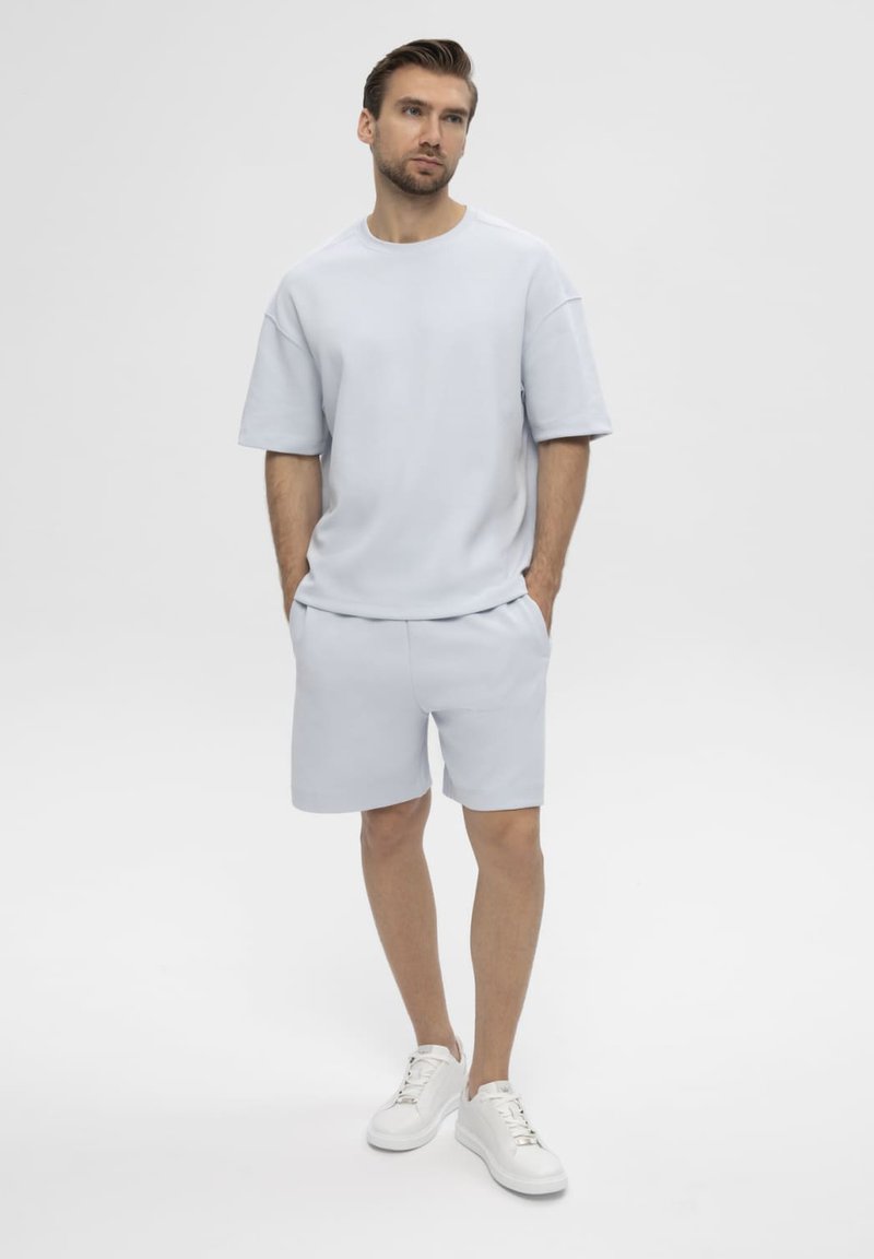 Jeune homme debout, les mains dans les poches, portant une chemise oversize bleu clair à manches courtes, un short assorti et des baskets blanches sur fond blanc.