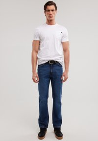 Uomo in piedi davanti a uno sfondo semplice, indossa una maglietta bianca, jeans blu con cintura nera e sneakers nere con suole marroni.