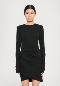 Vrouw met donker haar in een elegante zwarte lange mouw jurk met rimpeldetails en een asymmetrische zoom, staande tegen een effen lichte achtergrond.